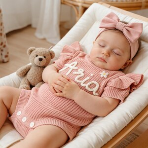 May include: A sleeping baby wearing a pink knitted romper with the name "Aria" embroidered in white. The romper has ruffled sleeves and a small daisy detail. A matching pink bow headband and a stuffed teddy bear are also visible.