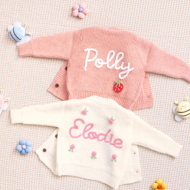 Peut inclure: Deux petits cardigans pour b&eacute;b&eacute;s, un rose et un cr&egrave;me, avec des noms brod&eacute;s et des d&eacute;tails floraux. Le cardigan rose porte l'inscription "Polly" en &eacute;criture blanche avec un &eacute;cusson fraise. Le cardigan cr&egrave;me porte l'inscription "Elodie" en &eacute;criture rose avec des motifs floraux.