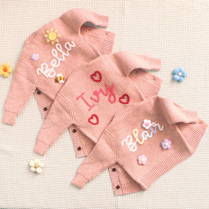 Peut inclure: Trois cardigans roses en tricot avec les noms brod&eacute;s : "Bella", "Ivy" et "Blair". Chaque pull pr&eacute;sente des accents floraux et en forme de c&oelig;ur d&eacute;coratifs. Les cardigans ont des fermetures &agrave; boutons et sont pr&eacute;sent&eacute;s avec des fleurs d&eacute;coratives.