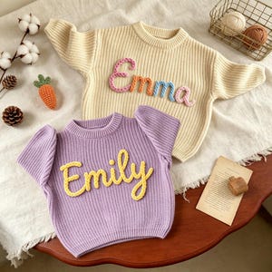 May include: A cream-colored long-sleeve sweater with the name Emma in multi-colored yarn lettering and a purple short-sleeve sweater with the name Emily in yellow yarn lettering, both featuring a ribbed knit texture and crew necklines.