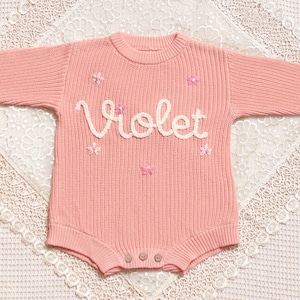 May include: A pale pink knitted baby romper with the name "Violet" in cream script across the chest. The romper has small pink flower appliqu&eacute;s and three wooden buttons at the bottom. The garment is displayed on a white lace surface.