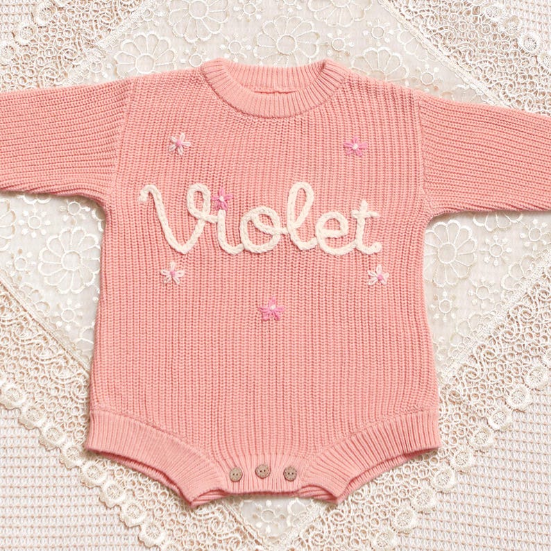 May include: A pale pink knitted baby romper with the name "Violet" in cream script across the chest. The romper has small pink flower appliqu&eacute;s and three wooden buttons at the bottom. The garment is displayed on a white lace surface.