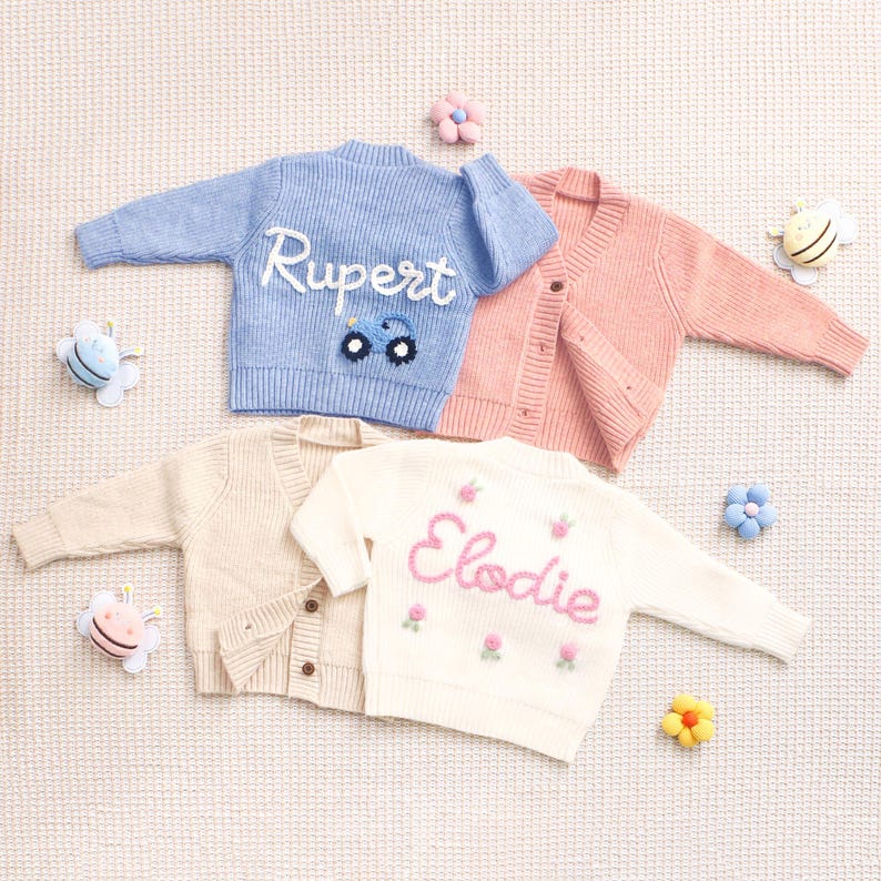 Peut inclure: Quatre cardigans en tricot de couleurs pastel. Un cardigan bleu clair avec le nom "Rupert" brod&eacute; en blanc et un motif de voiture. Un cardigan rose, un cardigan cr&egrave;me et un cardigan blanc avec le nom "Elodie" brod&eacute; en rose avec des motifs floraux.