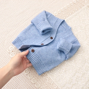 Peut inclure: Un cardigan en tricot bleu clair avec des poignets et un ourlet c&ocirc;tel&eacute;s. Le cardigan a un col en V et se ferme avec trois boutons marron. Le v&ecirc;tement est fait d'une mati&egrave;re douce et textur&eacute;e.