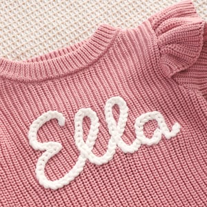 May include: A pink knitted sweater with the name "Ella" embroidered in white cursive script. The sweater has a ribbed texture and a ruffled sleeve detail. The background is a light beige, textured surface.