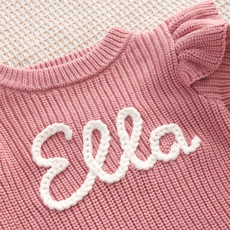 May include: A pink knitted sweater with the name "Ella" embroidered in white cursive script. The sweater has a ribbed texture and a ruffled sleeve detail. The background is a light beige, textured surface.