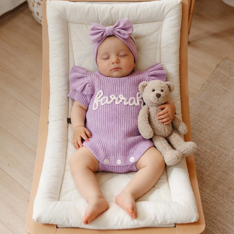 May include: A baby wearing a lavender knit romper with the name "Farrah" embroidered in white, a matching bow headband, and holding a beige teddy bear. The baby is lying on a white padded surface in a wooden rocker.