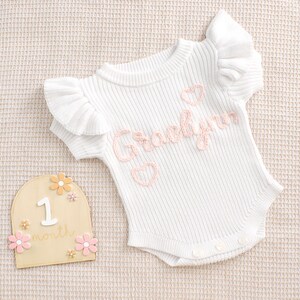 May include: White baby bodysuit with ruffled sleeves and the name "Gracelynn" embroidered in pink, along with pink hearts. A wooden milestone marker with the number "1" and the word "month" is also present, decorated with pink and orange flowers.