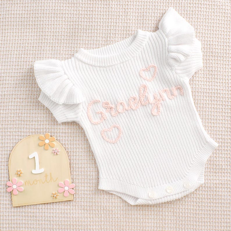 May include: White baby bodysuit with ruffled sleeves and the name "Gracelynn" embroidered in pink, along with pink hearts. A wooden milestone marker with the number "1" and the word "month" is also present, decorated with pink and orange flowers.