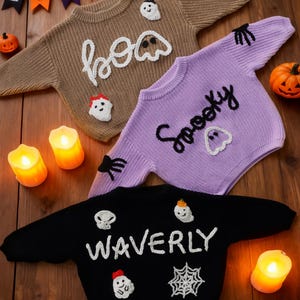Op de afbeelding: Drie Halloween-truien in bruin, paars en zwart. De bruine trui zegt "boo" met spookapplicaties. De paarse trui zegt "Spooky" met spookapplicaties. De zwarte trui zegt "WAVERLY" met schedel-, spook- en spinnenwebapplicaties.