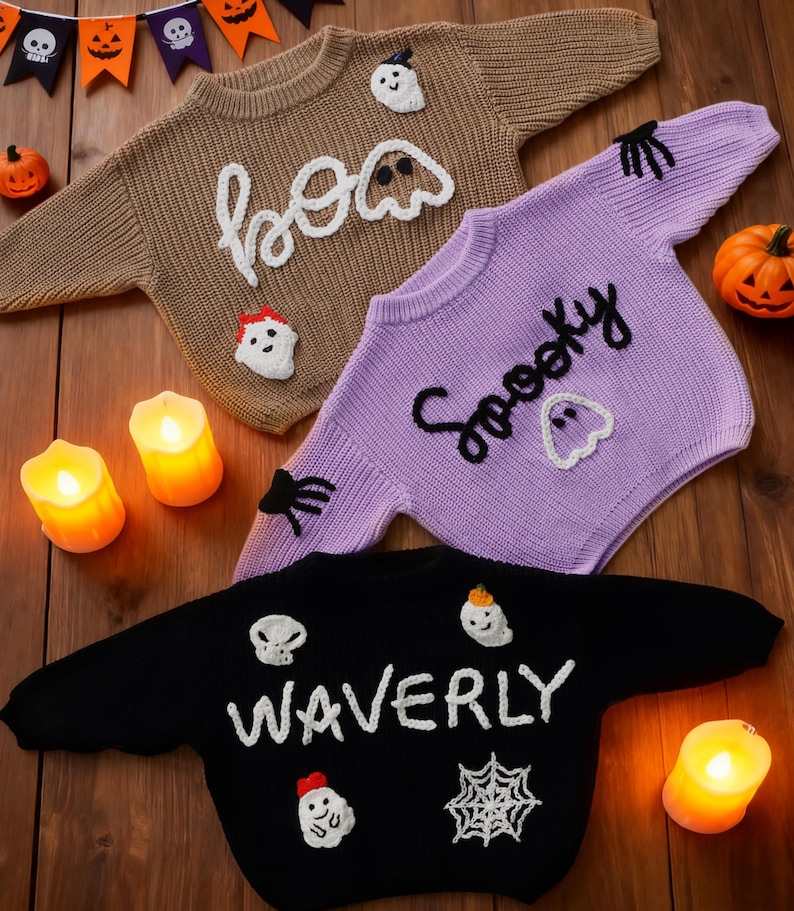 Op de afbeelding: Drie Halloween-truien in bruin, paars en zwart. De bruine trui zegt "boo" met spookapplicaties. De paarse trui zegt "Spooky" met spookapplicaties. De zwarte trui zegt "WAVERLY" met schedel-, spook- en spinnenwebapplicaties.