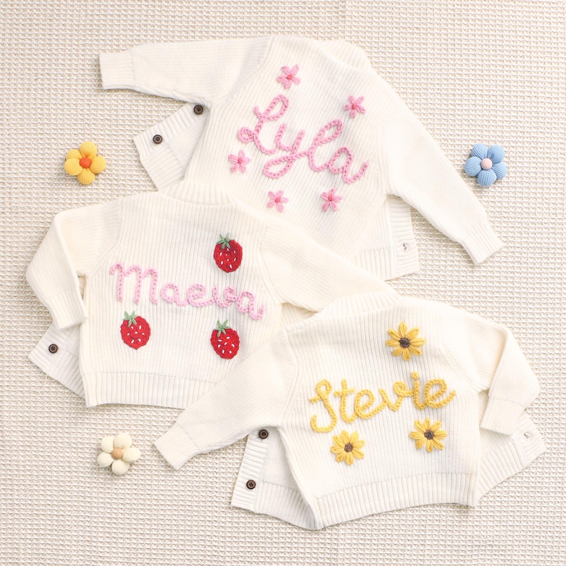 Peut inclure: Trois cardigans en tricot blanc cass&eacute; avec des noms et des motifs brod&eacute;s. L'un porte "Lyla" avec des fleurs roses, un autre "Maeve" avec des fraises, et le dernier "Stevie" avec des fleurs jaunes. Chaque cardigan a des boutons.