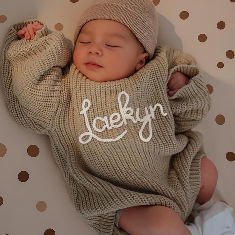May include: A sleeping baby wearing a beige knitted sweater and matching beanie. The sweater features the name "Laekyn" in white script. The baby is lying on a surface with a pattern of brown polka dots.