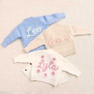 Peut inclure: Trois pulls pour b&eacute;b&eacute;s personnalis&eacute;s, bleu clair, beige et blanc. Le pull bleu porte le nom "Leo" en &eacute;criture blanche. Le pull beige porte "Gracie" en &eacute;criture rose. Le cardigan blanc porte "Lyla" en &eacute;criture rose avec des fleurs roses.