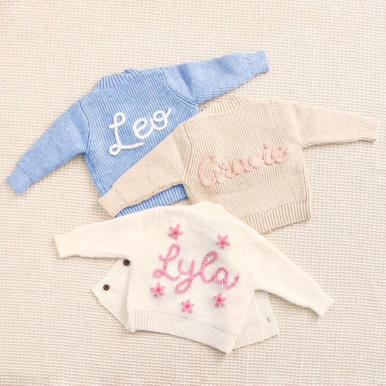 Peut inclure: Trois pulls pour b&eacute;b&eacute;s personnalis&eacute;s, bleu clair, beige et blanc. Le pull bleu porte le nom "Leo" en &eacute;criture blanche. Le pull beige porte "Gracie" en &eacute;criture rose. Le cardigan blanc porte "Lyla" en &eacute;criture rose avec des fleurs roses.