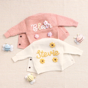 Peut inclure: Deux cardigans pour enfants, un rose et un cr&egrave;me, avec des noms brod&eacute;s et des d&eacute;tails floraux. Le rose dit "Blair" avec des fleurs blanches et violettes. Le cr&egrave;me dit "Stevie" avec des tournesols jaunes. Des jouets en forme d'abeilles sont dispos&eacute;s autour.