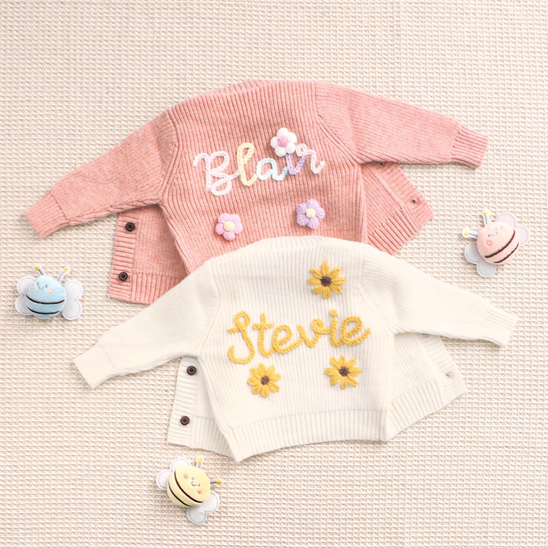 Peut inclure: Deux cardigans pour enfants, un rose et un cr&egrave;me, avec des noms brod&eacute;s et des d&eacute;tails floraux. Le rose dit "Blair" avec des fleurs blanches et violettes. Le cr&egrave;me dit "Stevie" avec des tournesols jaunes. Des jouets en forme d'abeilles sont dispos&eacute;s autour.