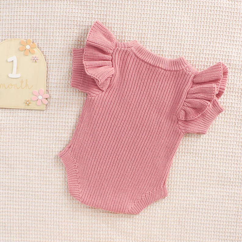 May include: A pink knitted baby bodysuit with ruffled sleeves. The one-piece garment is made of a ribbed knit fabric. A wooden sign with the number 1 and the word "month" is visible in the background.