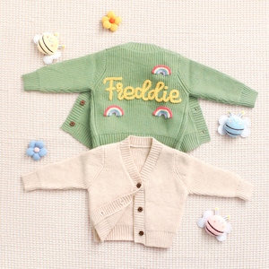 Peut inclure: Deux cardigans pour enfants. L'un est vert clair avec le nom "Freddie" en &eacute;criture jaune et des accents arc-en-ciel. L'autre est de couleur cr&egrave;me. Les deux ont des fermetures &agrave; boutons et des manches longues. Des jouets d&eacute;coratifs entourent les pulls.