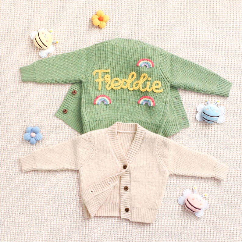Peut inclure: Deux cardigans pour enfants. L'un est vert clair avec le nom "Freddie" en &eacute;criture jaune et des accents arc-en-ciel. L'autre est de couleur cr&egrave;me. Les deux ont des fermetures &agrave; boutons et des manches longues. Des jouets d&eacute;coratifs entourent les pulls.