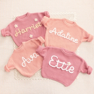 May include: Four long-sleeved knit sweaters in shades of pink and peach. Each sweater features a personalized name embroidered in white or yellow yarn across the chest. The names Harriet, Adaline, Ava, and Ettie are displayed, with small flower accents.
