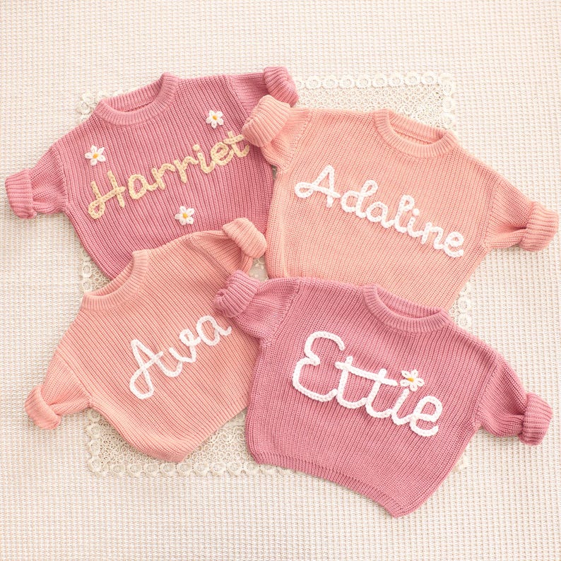 May include: Four long-sleeved knit sweaters in shades of pink and peach. Each sweater features a personalized name embroidered in white or yellow yarn across the chest. The names Harriet, Adaline, Ava, and Ettie are displayed, with small flower accents.
