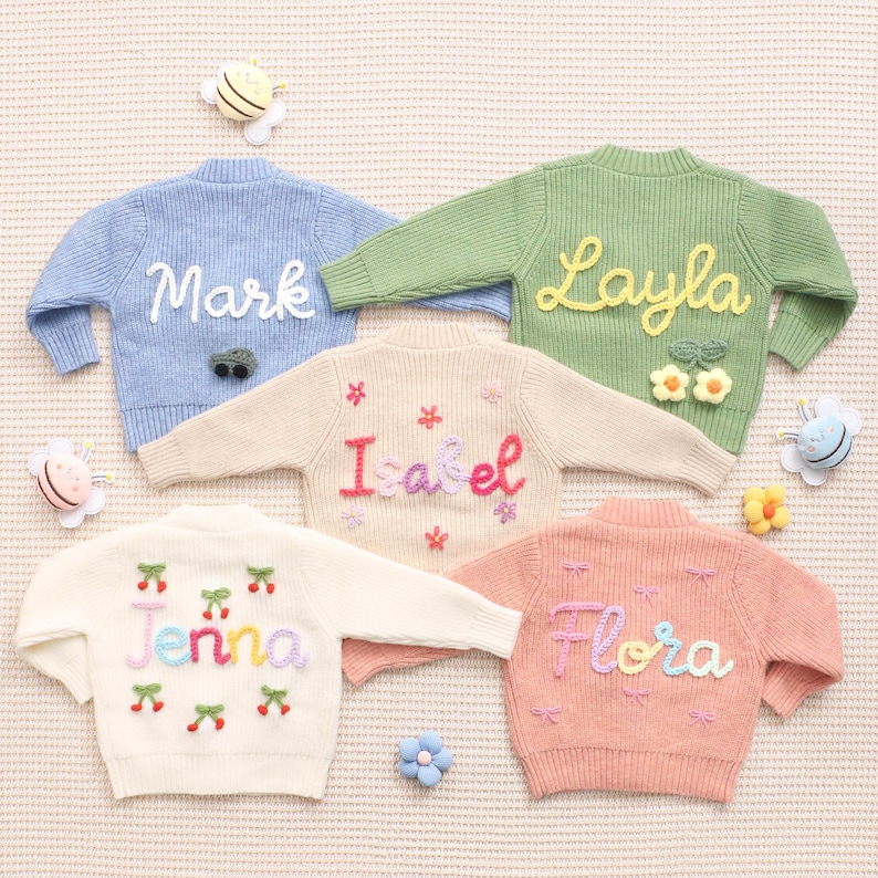 Peut inclure: Cinq pulls en tricot c&ocirc;tel&eacute; de couleurs pastel avec des noms personnalis&eacute;s brod&eacute;s sur le devant. Les pulls sont bleu clair, vert, beige, blanc et p&ecirc;che. Chaque pull porte un nom diff&eacute;rent dans une police color&eacute;e et fantaisiste. &Eacute;l&eacute;ments d&eacute;coratifs : fleurs, cerises et n&oelig;uds.