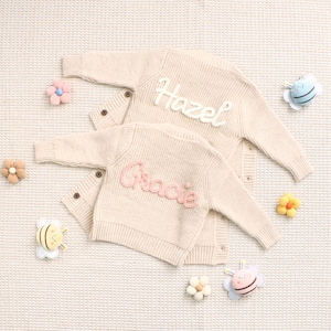 Peut inclure: Deux cardigans en tricot cr&egrave;me personnalis&eacute;s, l'un avec "Hazel" en blanc et l'autre avec "Gracie" en rose. Des d&eacute;corations florales et en forme d'abeilles entourent les pulls.