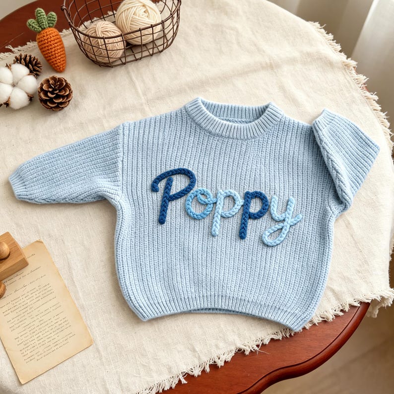 May include: A light blue long-sleeve knit sweater featuring the word Poppy embroidered in a gradient of blue yarn across the chest. The sweater has a ribbed crew neckline, cuffs, and hem, with a thick, textured knit construction throughout.