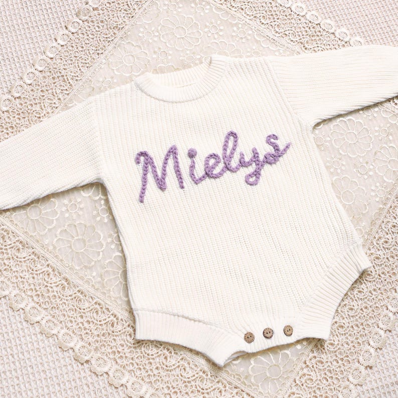 May include: A white knitted baby romper with the name "Mielys" embroidered in purple script across the chest. The romper has long sleeves and three wooden buttons at the bottom. The garment is displayed on a white lace surface.