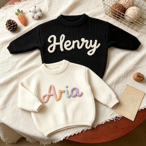 May include: A black long-sleeve sweater with the name Henry in white yarn lettering and a cream long-sleeve sweater with the name Aria in multicolored yarn lettering. Both sweaters feature a ribbed knit texture and a crew neckline design.