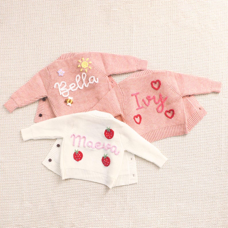 Peut inclure: Trois petits cardigans pour b&eacute;b&eacute;s, roses et blancs. Un cardigan rose porte le nom "Bella" brod&eacute; avec un soleil et une abeille. Un autre rose porte "Ivy" avec des c&oelig;urs. Le cardigan blanc porte "Maeve" avec des fraises.