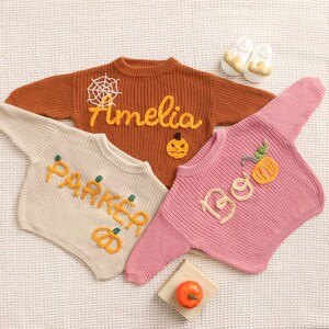 Op de afbeelding: Drie gebreide baby truien in oranje, cr&egrave;me en roze, elk met geborduurde Halloween-thema ontwerpen. De oranje trui heeft een spinnenweb en de naam "Amelia". De cr&egrave;mekleurige trui zegt "Parker". De roze trui zegt "Boo".