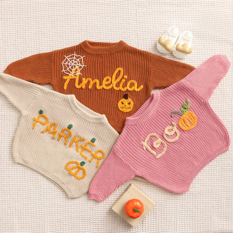 Op de afbeelding: Drie gebreide baby truien in oranje, cr&egrave;me en roze, elk met geborduurde Halloween-thema ontwerpen. De oranje trui heeft een spinnenweb en de naam "Amelia". De cr&egrave;mekleurige trui zegt "Parker". De roze trui zegt "Boo".