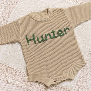 May include: A beige knitted baby romper with long sleeves. The word "Hunter" is embroidered in green across the chest. The romper has a round neckline and three buttons at the bottom. The garment is displayed on a white lace surface.