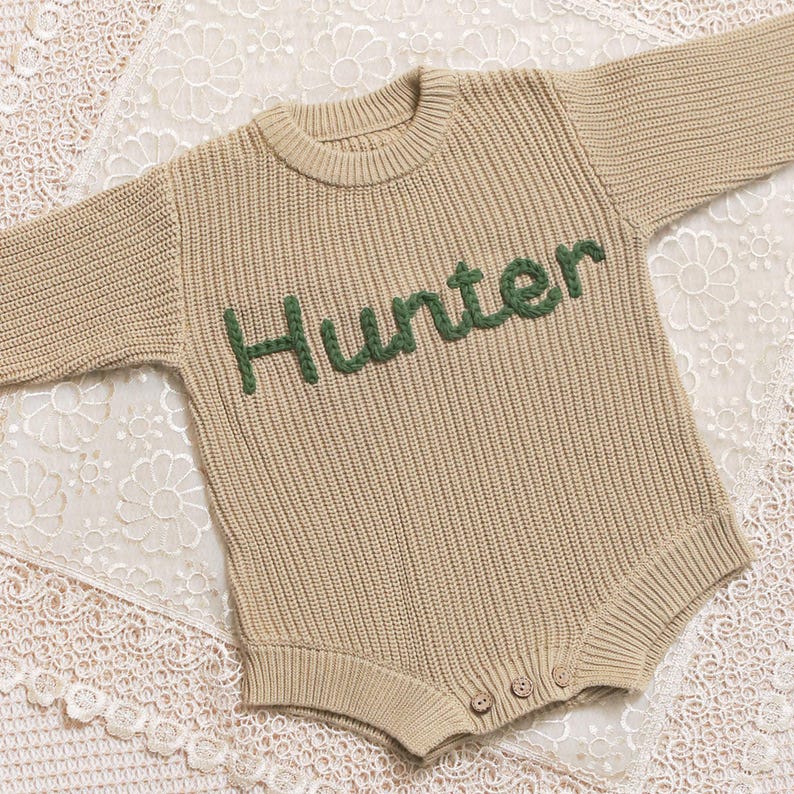 May include: A beige knitted baby romper with long sleeves. The word "Hunter" is embroidered in green across the chest. The romper has a round neckline and three buttons at the bottom. The garment is displayed on a white lace surface.