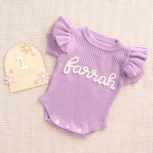 May include: A lilac-colored baby romper with ruffled sleeves and the name "Farrah" in white script. The romper has three buttons at the bottom. A wooden milestone marker with the number "1" and the word "month" is also present.