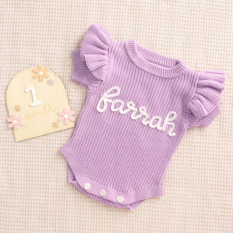 May include: A lilac-colored baby romper with ruffled sleeves and the name "Farrah" in white script. The romper has three buttons at the bottom. A wooden milestone marker with the number "1" and the word "month" is also present.