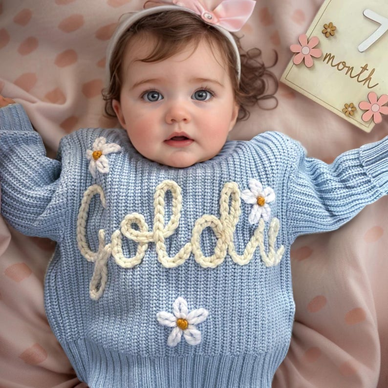 May include: A light blue knit sweater featuring the name Goldie in cream-colored yarn lettering across the chest, accented with three white and yellow embroidered daisy flowers. The sweater has a ribbed texture and long sleeves.