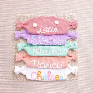 May include: A stack of five long-sleeve knit sweaters in pink, lavender, mint green, peach, and white. Each sweater features a name embroidered in cursive yarn: Little, Eloise, Meira, Nancu, and Chelsea, with small flower accents on some.