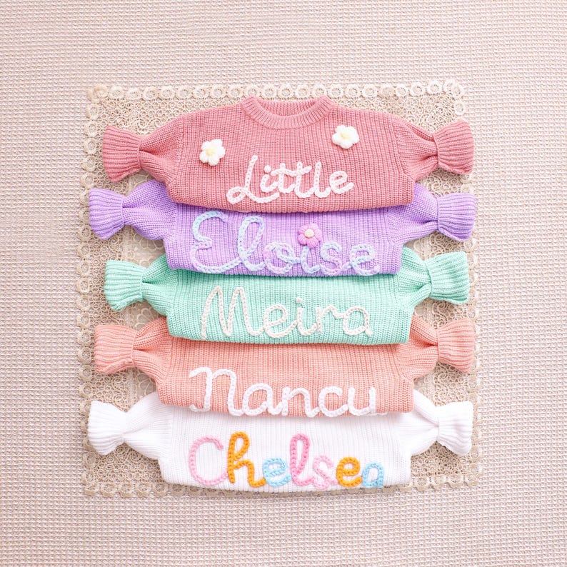 May include: A stack of five long-sleeve knit sweaters in pink, lavender, mint green, peach, and white. Each sweater features a name embroidered in cursive yarn: Little, Eloise, Meira, Nancu, and Chelsea, with small flower accents on some.