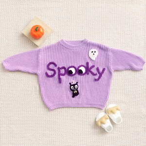 Op de afbeelding: Een lila gebreide trui met het woord "Spooky" in paars garen. De trui heeft een witte geest en een zwarte kat applicatie. Een kleine oranje pompoen ligt op een houten vierkant. Twee paar witte en gele babyschoenen staan ook op de foto.