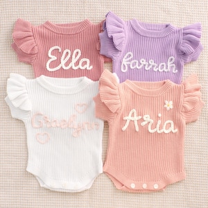 May include: Four baby bodysuits in pink, purple, white, and peach. Each has a name embroidered in white script: Ella, Farrah, Gracelyn, and Aria. The bodysuits have ruffled sleeves and a ribbed knit texture.
