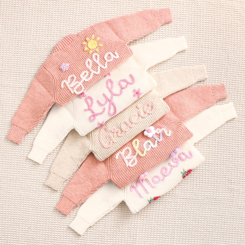 Peut inclure: Plusieurs pulls de couleurs pastel personnalis&eacute;s, chacun brod&eacute; d'un pr&eacute;nom en &eacute;criture cursive. Les pulls pr&eacute;sentent des ornements floraux et solaires, cr&eacute;ant un design charmant et unique.