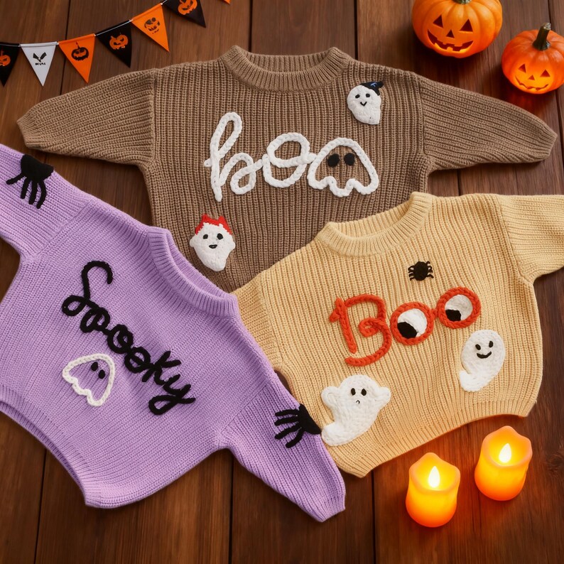Op de afbeelding: Drie Halloween-truien in bruin, geel en paars. De bruine trui zegt "boo" in het wit, de gele trui zegt "Boo" in het oranje en de paarse trui zegt "Spooky" in het zwart. Elke trui heeft spook- en spinnenapplicaties.