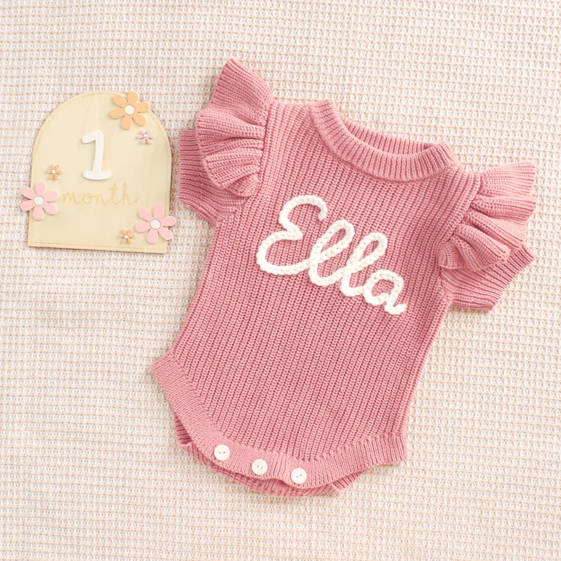 May include: A pink knitted baby romper with ruffled sleeves and the name "Ella" in white script. The romper has three white buttons at the bottom. A wooden milestone marker with the number "1" and the word "month" is also present.