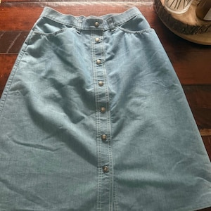 May include: A light blue denim skirt featuring a high-waisted design with a button-down front closure. The skirt includes two side pockets, a structured waistband with belt loops, and a vertical seam running down the center front panel.