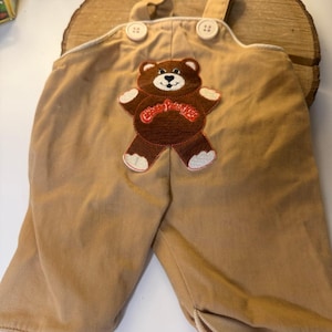 Puede incluir: Peto de algodón color tostado con dos botones claros en los tirantes y un parche central bordado de un oso marrón. El oso tiene detalles blancos en las patas y orejas, con el texto rojo Cabbage Patch Kids bordado sobre el pecho.