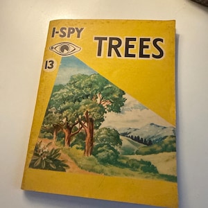 Könnte beinhalten: Ein Vintage-Buch "I-SPY TREES" mit gelbem Einband. Der Einband zeigt den Titel in großen schwarzen Buchstaben und eine Landschaftsillustration von Bäumen, Hügeln und einem Pfad. Das Buch ist leicht abgenutzt.