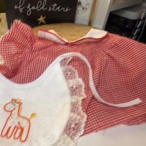 May include: A red and white gingham dress with a white collar and a white bib featuring a lace trim and an embroidered orange giraffe. The dress has short sleeves and a rounded neckline, while the bib is secured with white fabric ties.